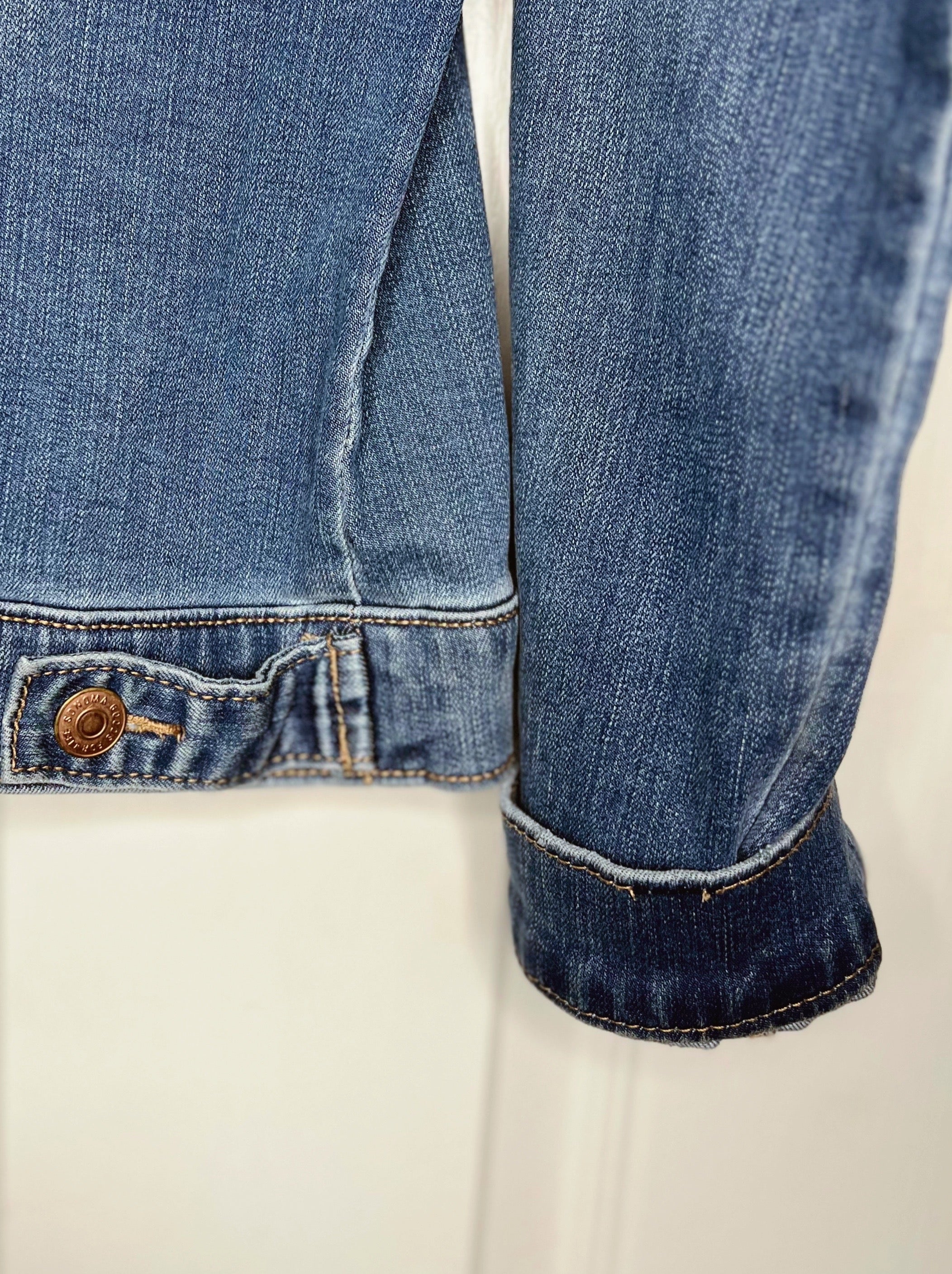 Close-up of a blue denim jacket sleeve with a rolled cuff and a buttoned pocket detail, shown against a plain light background.