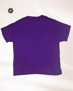 A solid purple short-sleeve t-shirt shown from the back, laid flat against a light off-white background. A black and white compass logo is visible in the top-left corner.