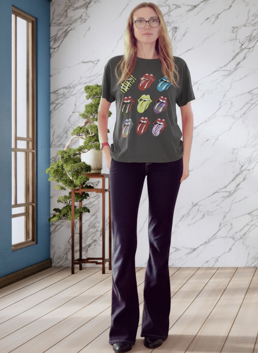 A woman wearing a dark gray Rolling Stones graphic t-shirt and dark flared jeans, standing in a room with a marble wall, wood floor, and a potted plant.