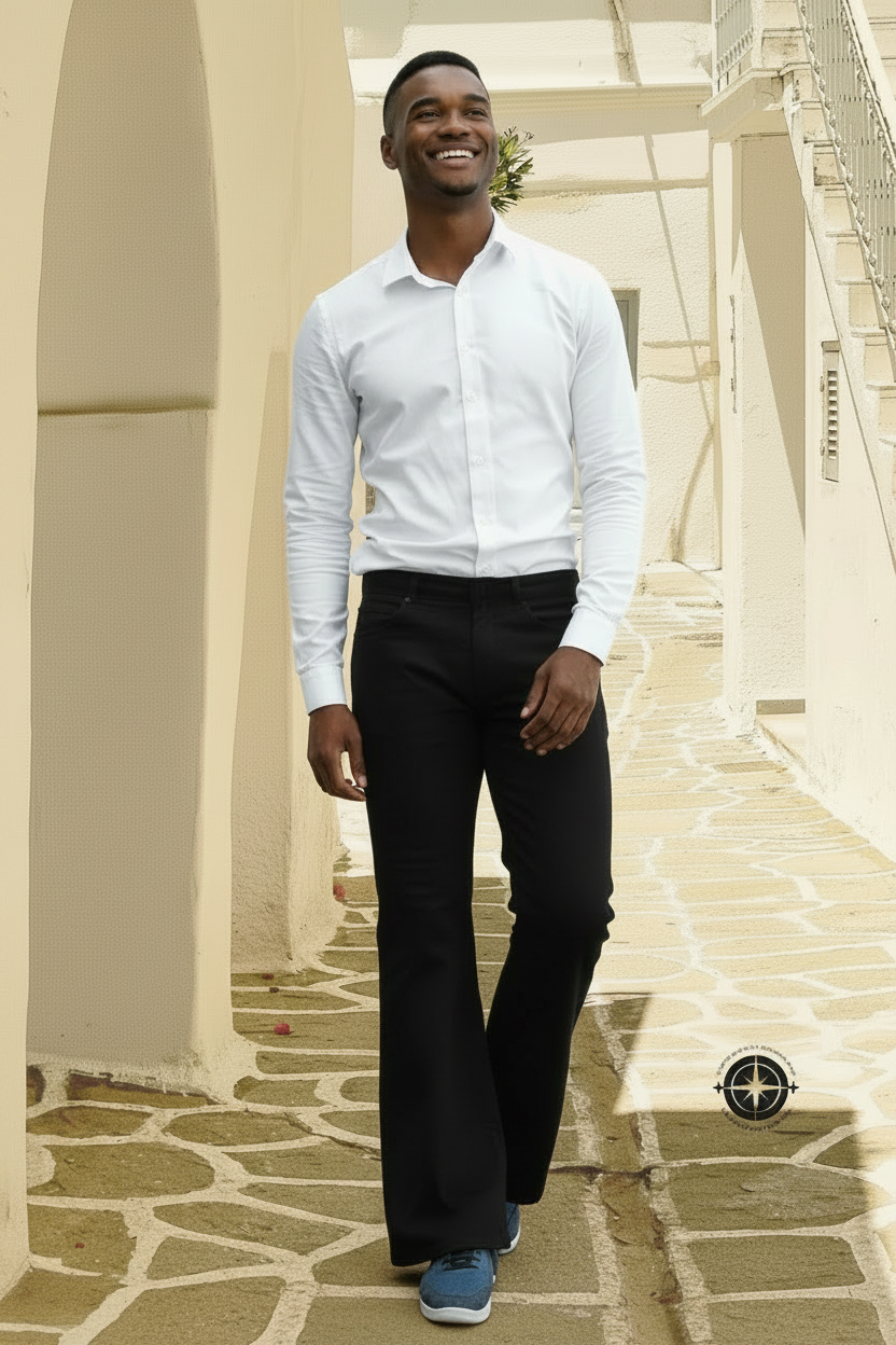 A man wearing a white long-sleeve button-up shirt, black flared trousers, and blue sneakers walking down a sunlit stone corridor with cream-colored arches. A compass logo is visible in the lower right corner.