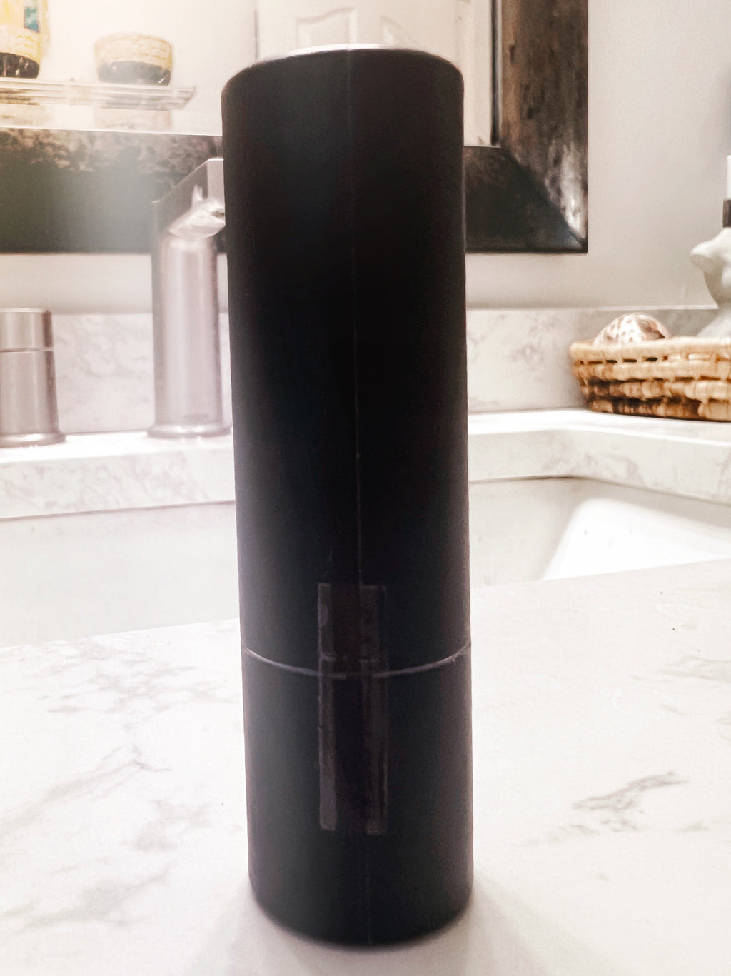 A matte black cylindrical container or bottle standing upright on a white marble bathroom counter with a sink and mirror in the background.