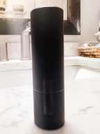 A matte black cylindrical container or bottle standing upright on a white marble bathroom counter with a sink and mirror in the background.
