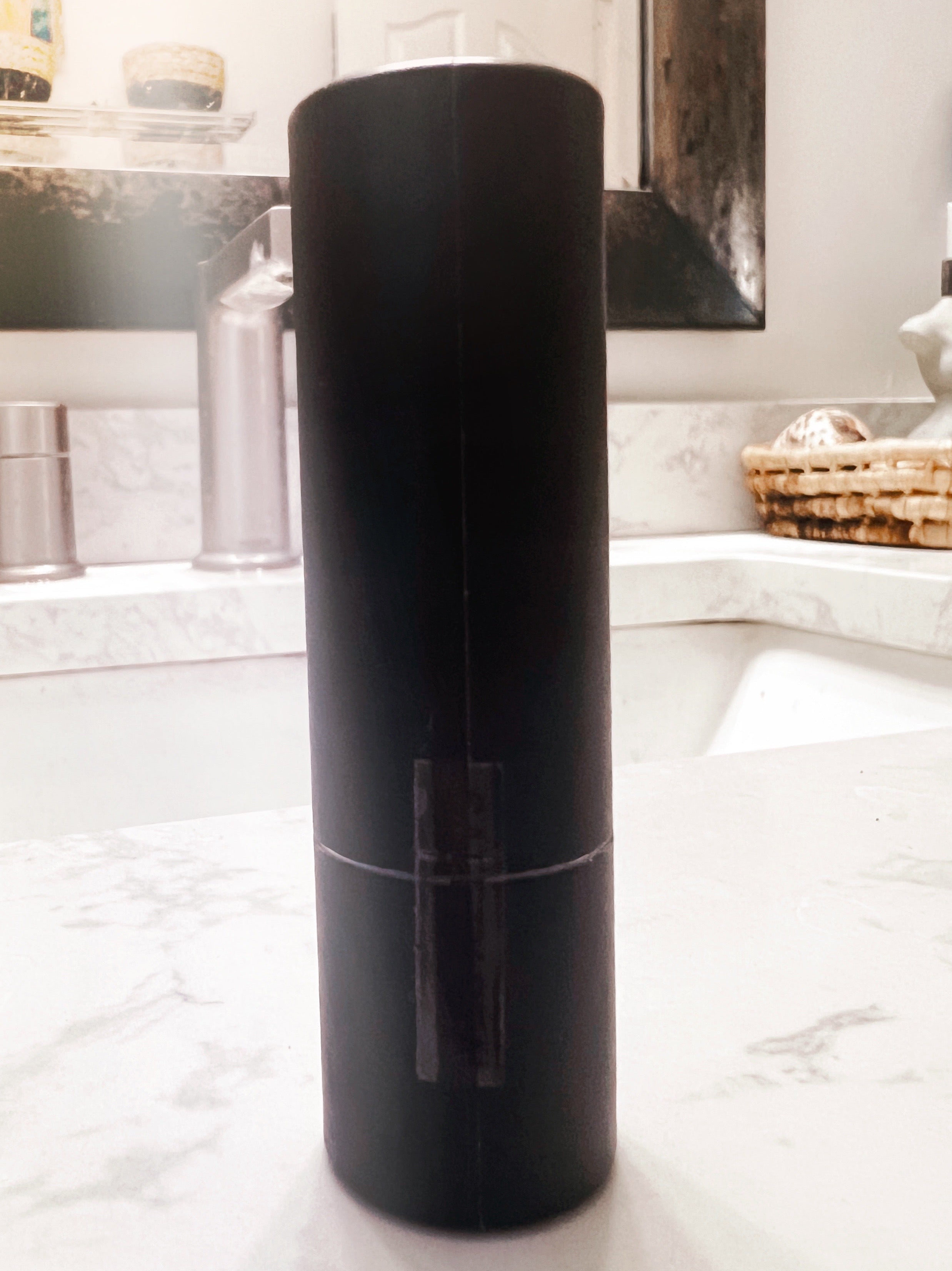 A matte black cylindrical container or bottle standing upright on a white marble bathroom counter with a sink and mirror in the background.