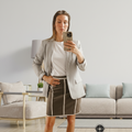 A woman takes a mirror selfie in a living room, wearing a blue graphic t-shirt and a brown pencil skirt with white piping. A compass logo is visible in the bottom right corner.