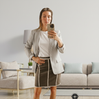 A woman takes a mirror selfie in a living room, wearing a blue graphic t-shirt and a brown pencil skirt with white piping. A compass logo is visible in the bottom right corner.