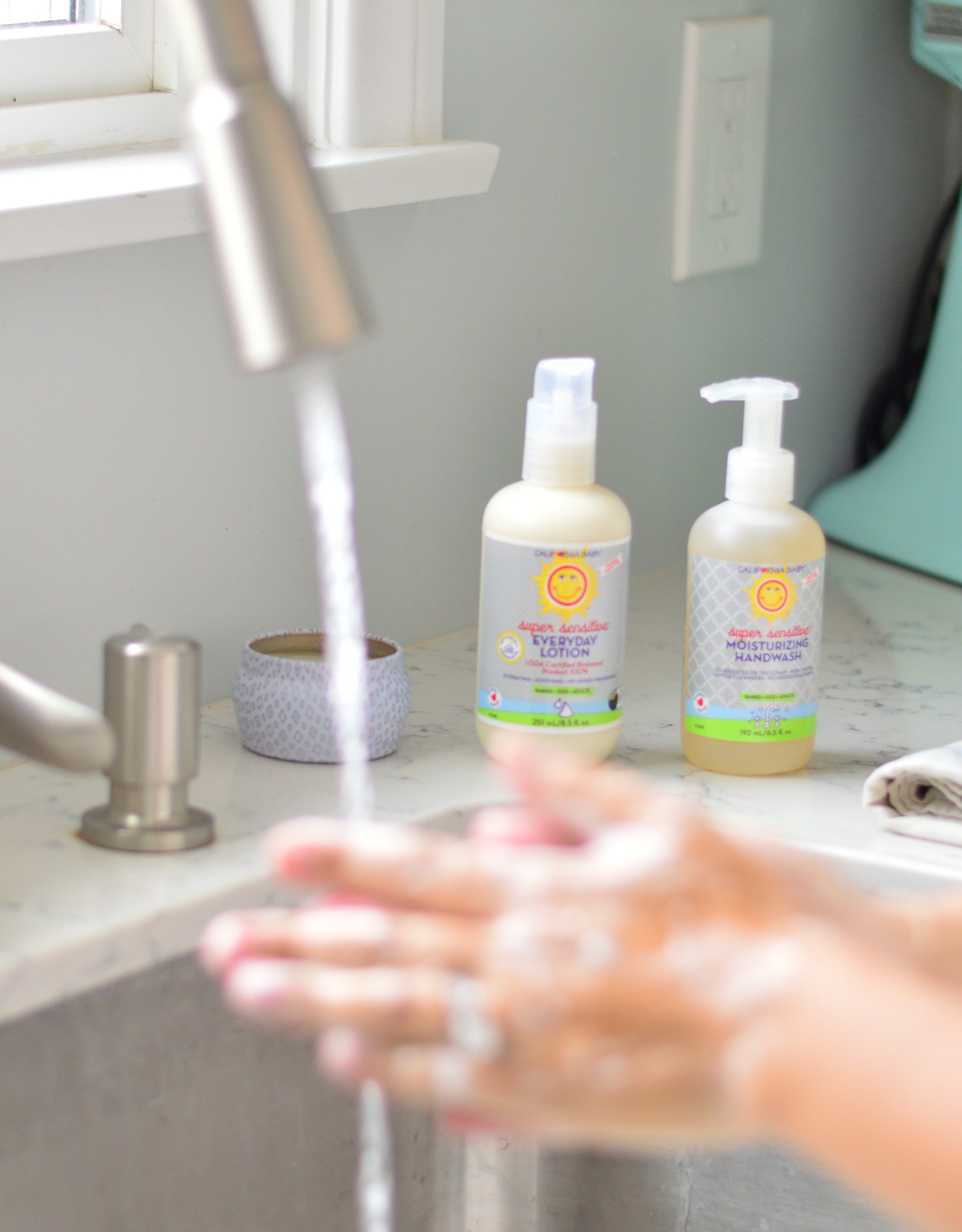 Person washing hands with soap under running water, with two bottles of hand sanitizer on a counter.