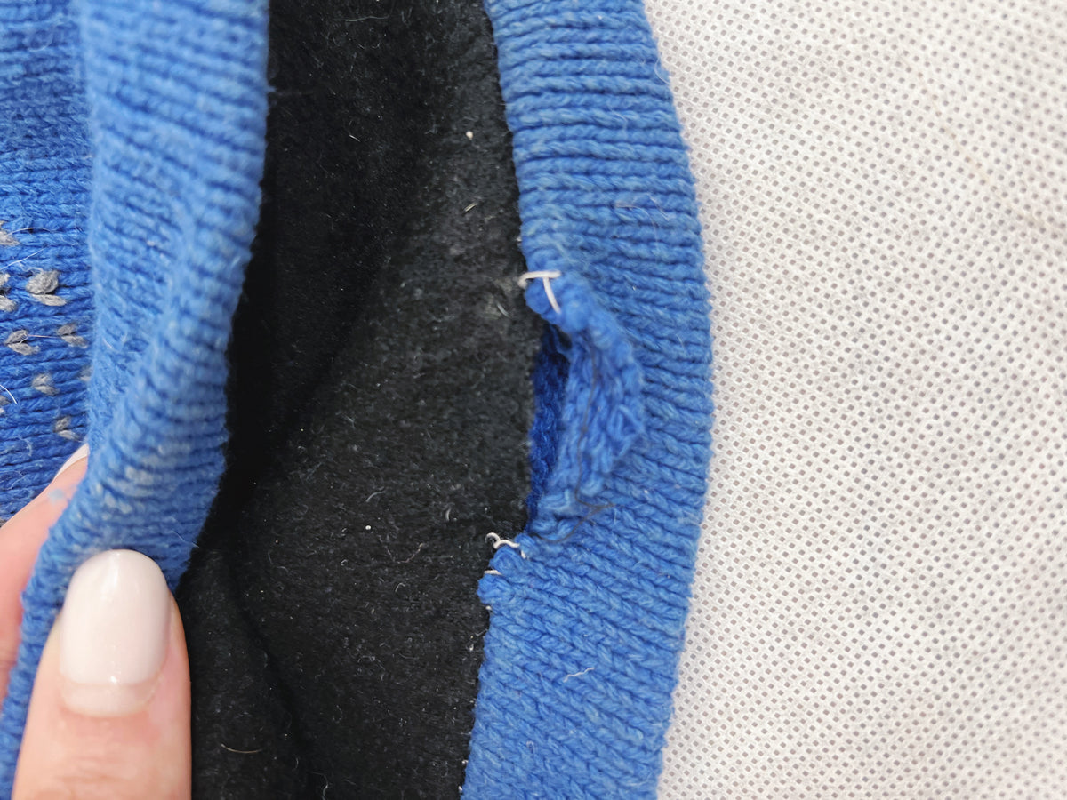 Close-up of a blue knit garment being held open by a hand, showing a black fleece or wool lining with a small loose thread detail, next to a white textured fabric background.