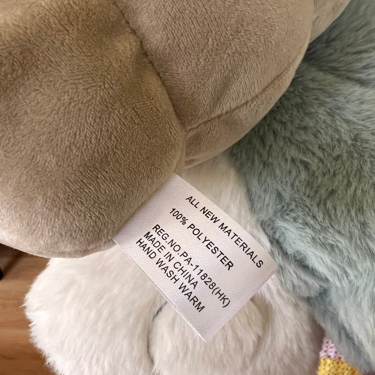 Close-up of a plush toy's fabric tag reading 'ALL NEW MATERIALS 100% POLYESTER REG NO.PA-11928(HK) MADE IN CHINA HAND WASH WARM' attached to beige, white, and pale blue plush fabric.