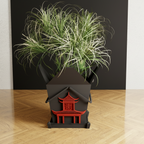 A black house-shaped planter with a red pagoda design, holding a green ornamental grass plant, set on a light wood floor against a dark background.