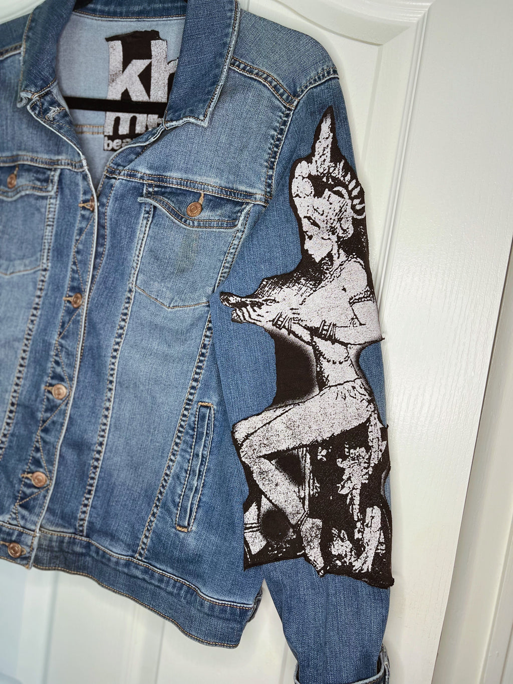 A blue denim jacket with a large black and white graphic patch of a pin-up style woman on the right sleeve, hanging against a white door.