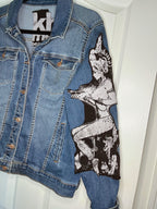A blue denim jacket with a large black and white graphic patch of a pin-up style woman on the right sleeve, hanging against a white door.