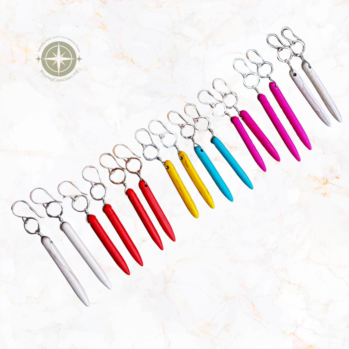 Six pairs of nail scissors arranged diagonally on a light marble background, showing color options from silver and white to red, rose gold, yellow, turquoise, and magenta. Each pair has a silver metal ring clasp and a long, tapered nail file shaped like a pin.
