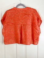 An orange short-sleeve blouse with a small floral print, hanging on a gray hanger against a white paneled door.