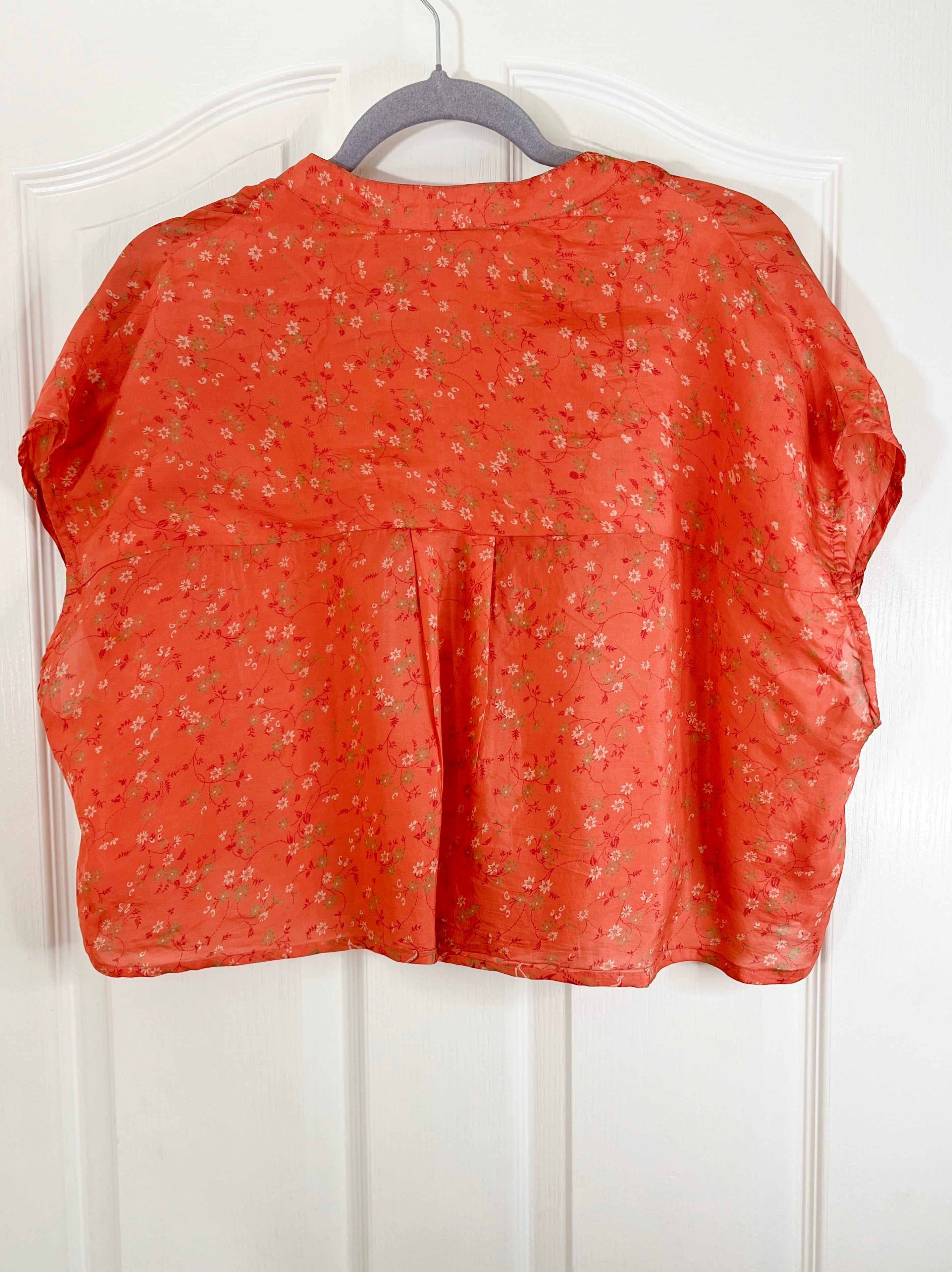 An orange short-sleeve blouse with a small floral print, hanging on a gray hanger against a white paneled door.