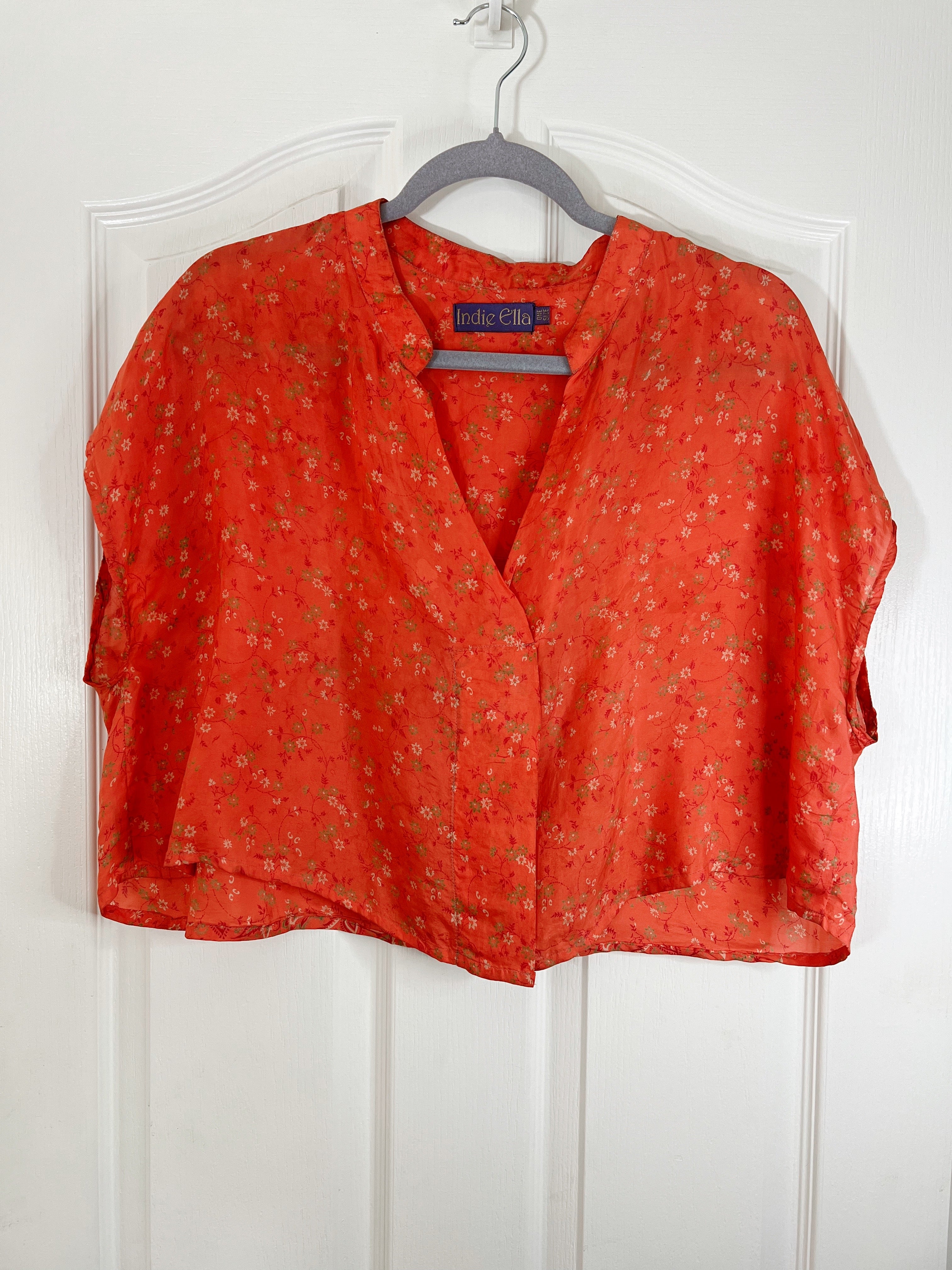 An orange-red short-sleeve blouse with a small floral print, hanging on a gray hanger against a white paneled door. The brand label inside the collar reads 'Majestic'.