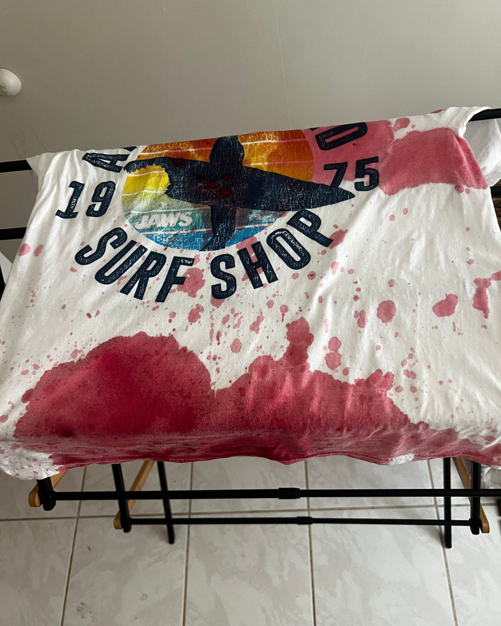 A white vintage-style surf shop t-shirt with a colorful graphic and '1975 SURF SHOP' text, heavily splattered with pink and red paint or ink, draped over a black metal stand.