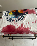 A white vintage-style surf shop t-shirt with a colorful graphic and '1975 SURF SHOP' text, heavily splattered with pink and red paint or ink, draped over a black metal stand.