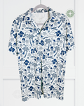 A white short-sleeve camp collar shirt with a blue all-over retro-style print featuring palm trees, surfboards, hibiscus flowers, and illustrated faces. The shirt is shown hanging on a hanger against a white paneled wall.