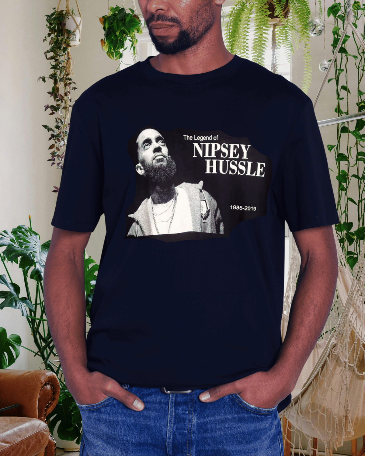 A man wearing a navy blue graphic t-shirt featuring a black and white photo of Nipsey Hussle with the text 'The Legend of NIPSEY HUSSLE 1985-2019'. The background is a cozy indoor setting with hanging plants and a hammock.