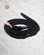 A coiled black braided rope with a metallic rose gold clasp, displayed on a textured light beige sand-like surface.