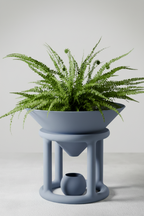 A modern blue-gray ceramic planter with a wide bowl-shaped top holding a green fern, supported by a sculptural multi-tiered stand with a small matching pot inside, set against a neutral light gray background.