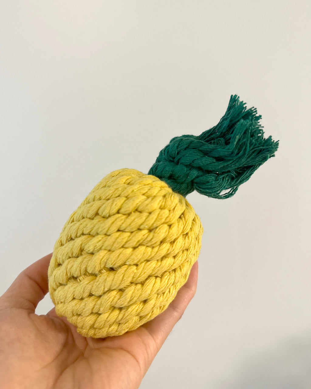Hand holding a yellow rope pineapple toy with a green top against a plain background