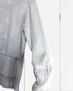A close-up of a sheer white embroidered blouse with intricate floral and geometric patterns, featuring a smocked sleeve and lace-trimmed hem.
