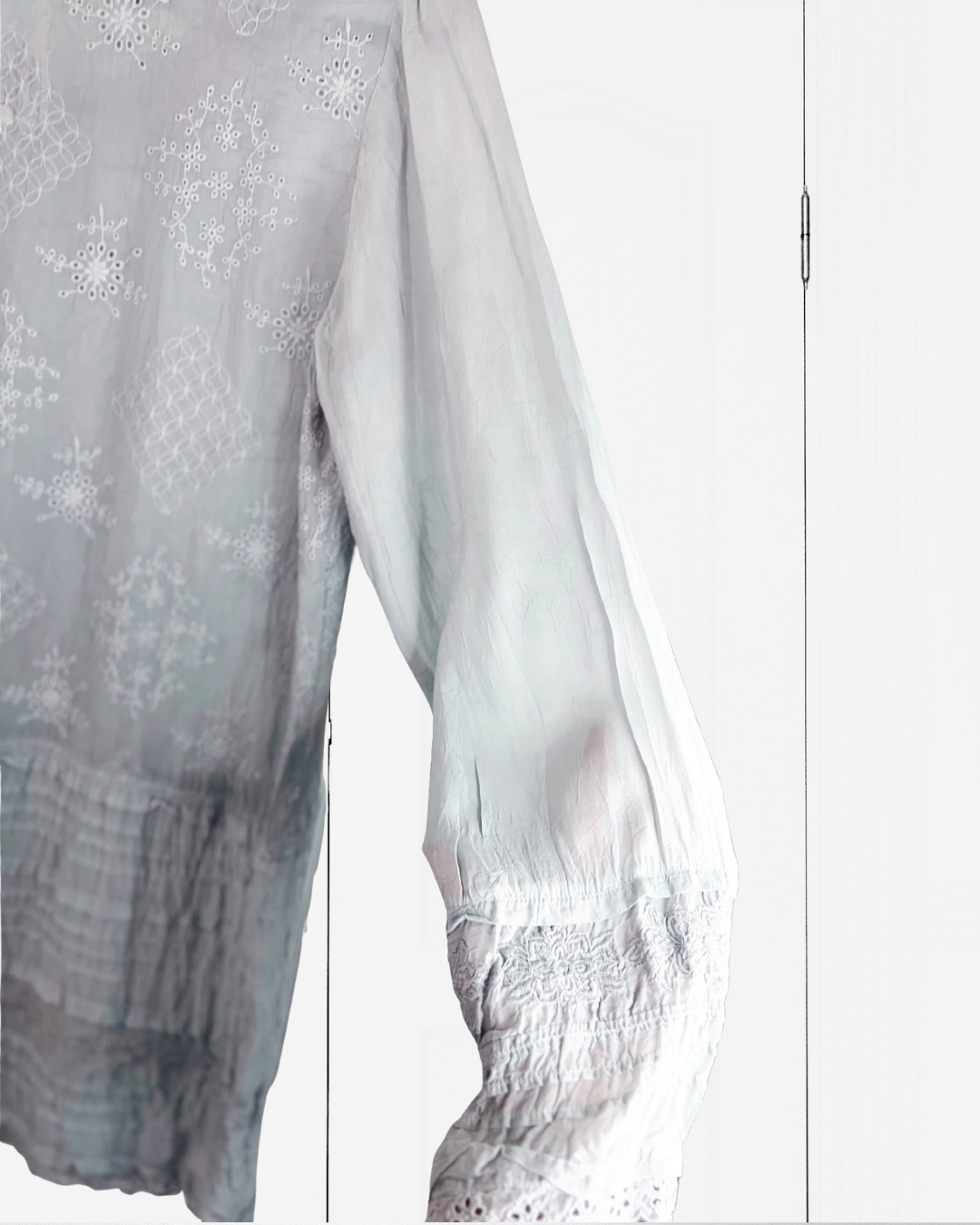 A close-up of a sheer white embroidered blouse with intricate floral and geometric patterns, featuring a smocked sleeve and lace-trimmed hem.