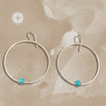 A pair of large silver-tone hoop earrings with a small round turquoise-blue stone at the bottom of each hoop, displayed on a textured beige background.