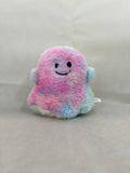 A small plush ghost toy with a pastel rainbow color scheme of pink, blue, and purple, featuring a simple black embroidered smiley face, set against a neutral gray fabric background.