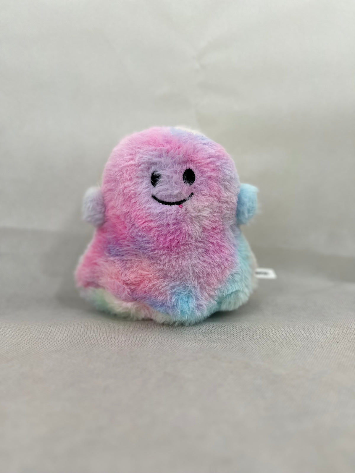 A small plush ghost toy with a pastel rainbow color scheme of pink, blue, and purple, featuring a simple black embroidered smiley face, set against a neutral gray fabric background.