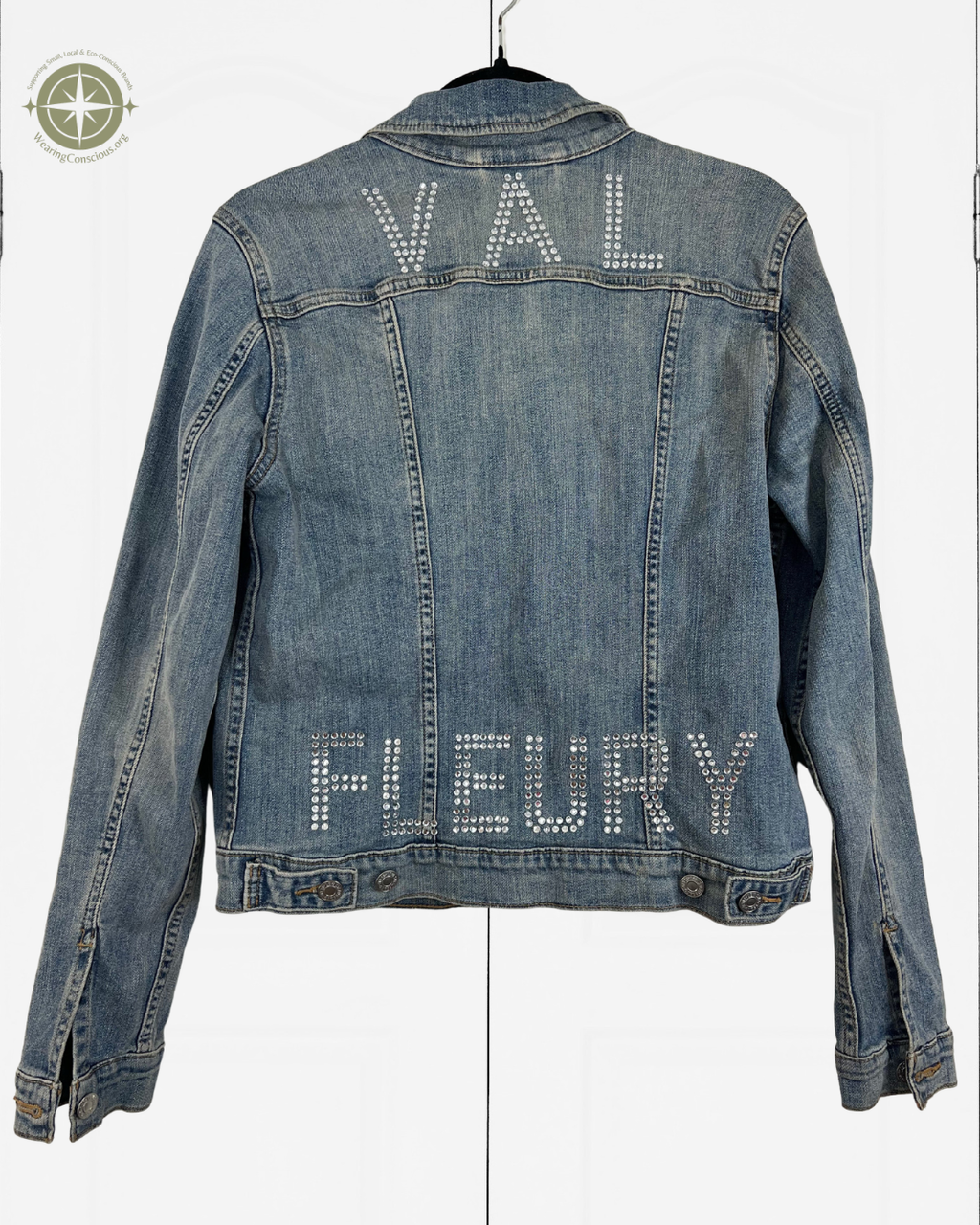 A blue denim jacket hanging on a black hanger against a white background, featuring the names 'VAL' and 'FLEURY' spelled out in rhinestones across the back.