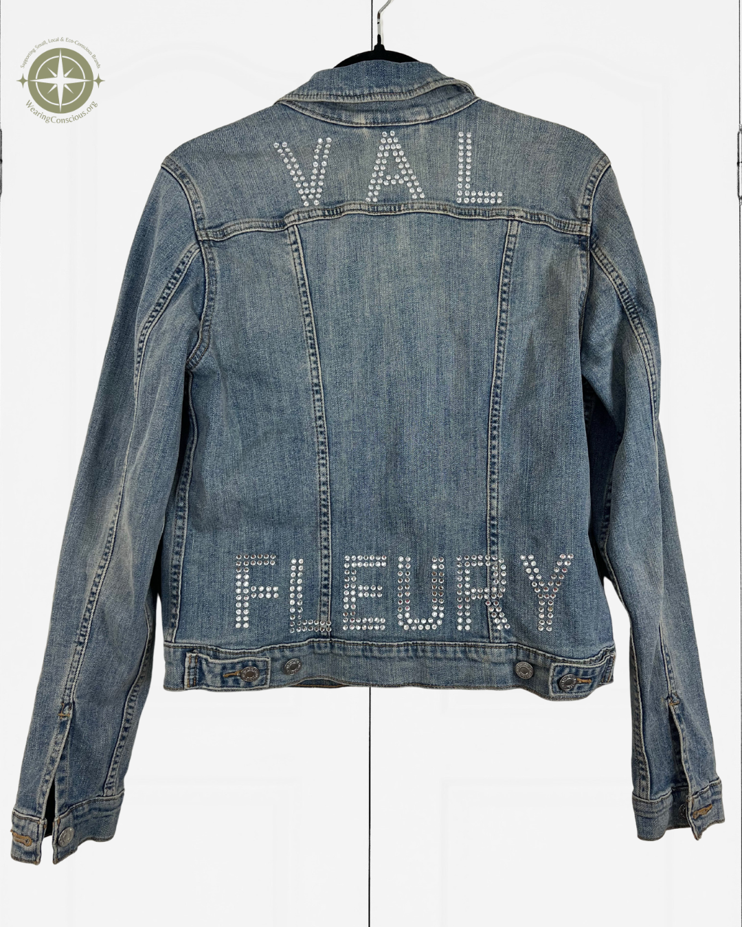 A blue denim jacket hanging on a black hanger against a white background, featuring the names 'VAL' and 'FLEURY' spelled out in rhinestones across the back.