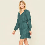 A woman modeling a dark teal green long-sleeve mini dress with a textured grid pattern, ruffled shoulders, and a tiered skirt, set against a plain white background.