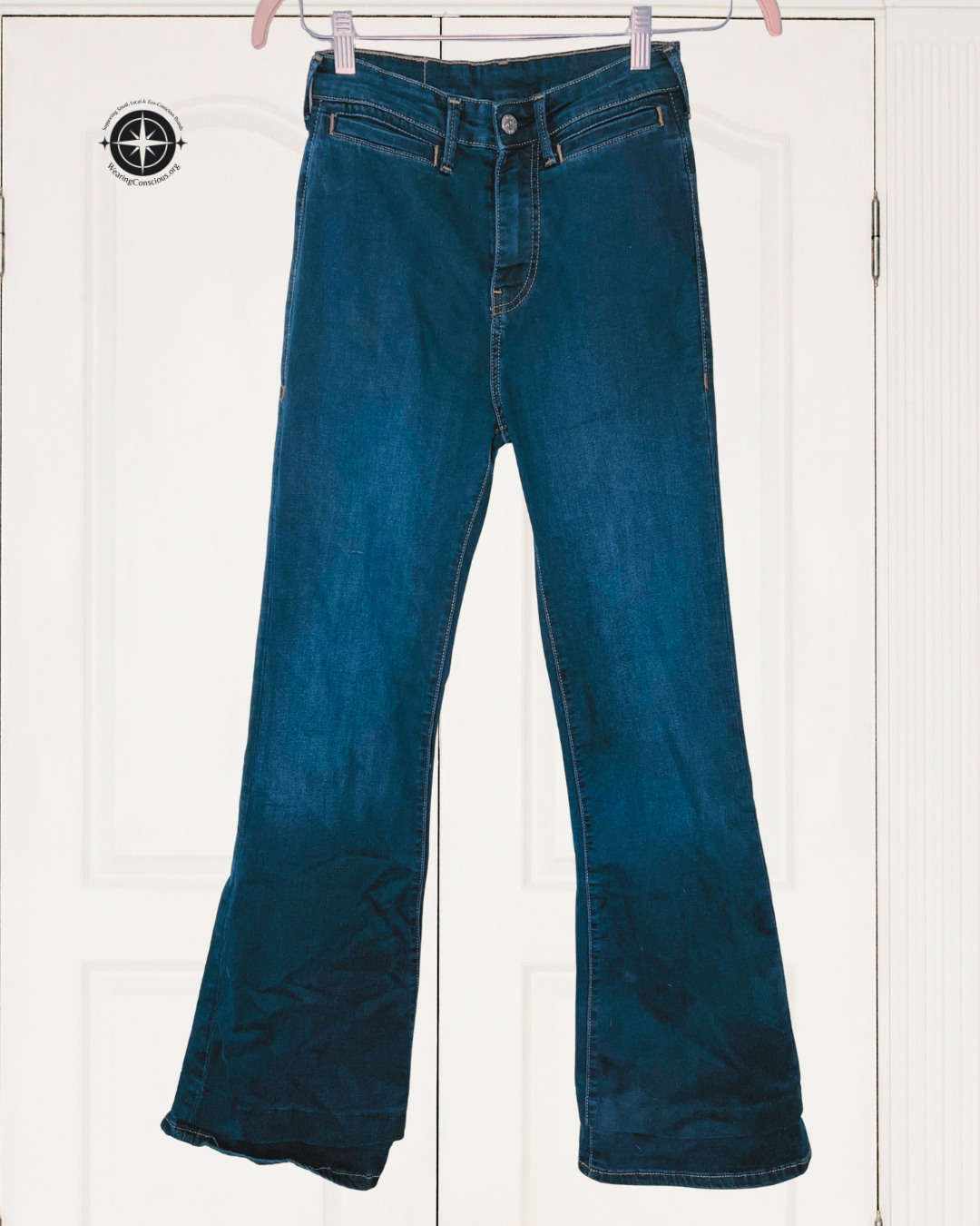 A pair of dark blue flare jeans hanging on pink hangers against a white paneled door background.