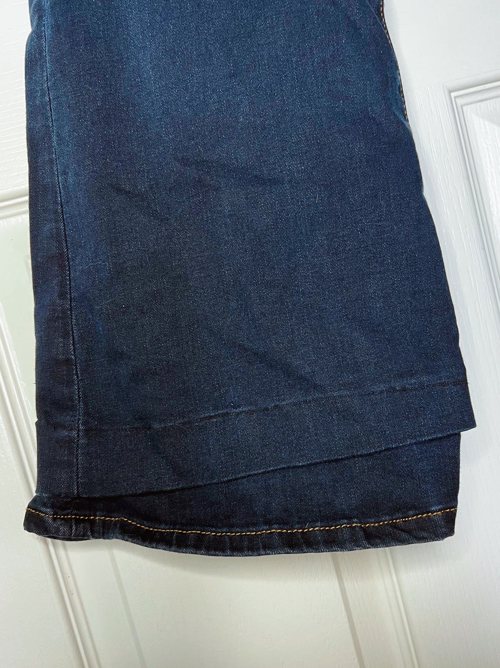 A close-up of a dark blue denim garment, likely jeans or trousers, folded over a white paneled door. The fabric shows a classic indigo wash with visible orange-tan contrast stitching along the hem.