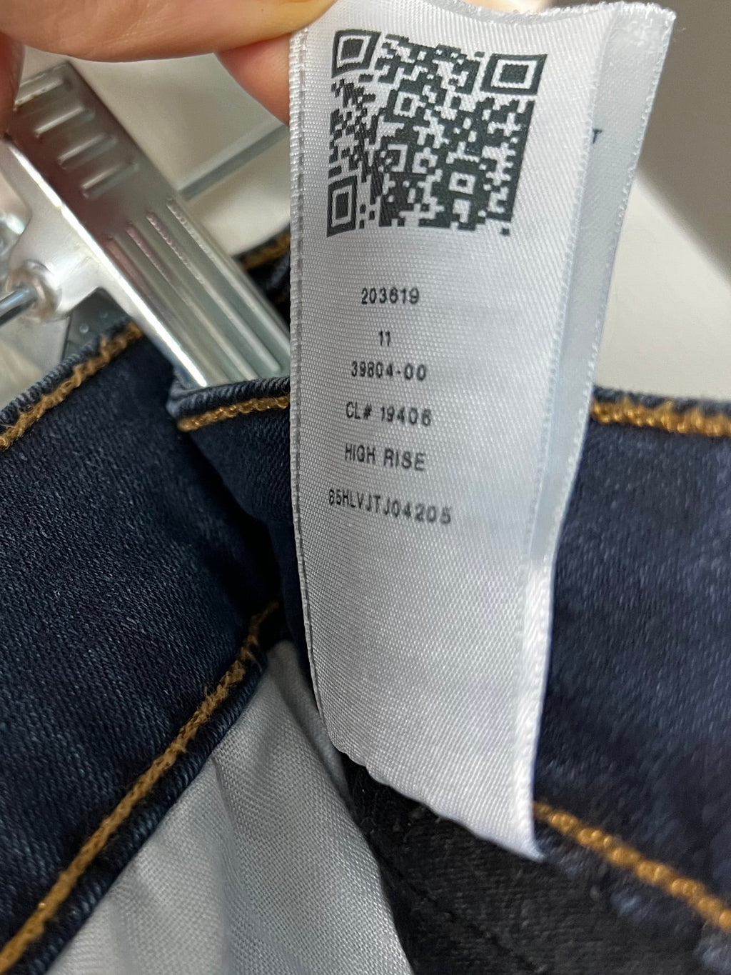Close-up of a denim garment label attached to dark blue jeans, showing a QR code and printed size information. A finger is holding the label, and the jeans are photographed against a light neutral background.