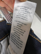 A close-up of a white fabric care label sewn onto dark blue denim jeans, held by a hand. The label contains German and English instructions for washing and care.