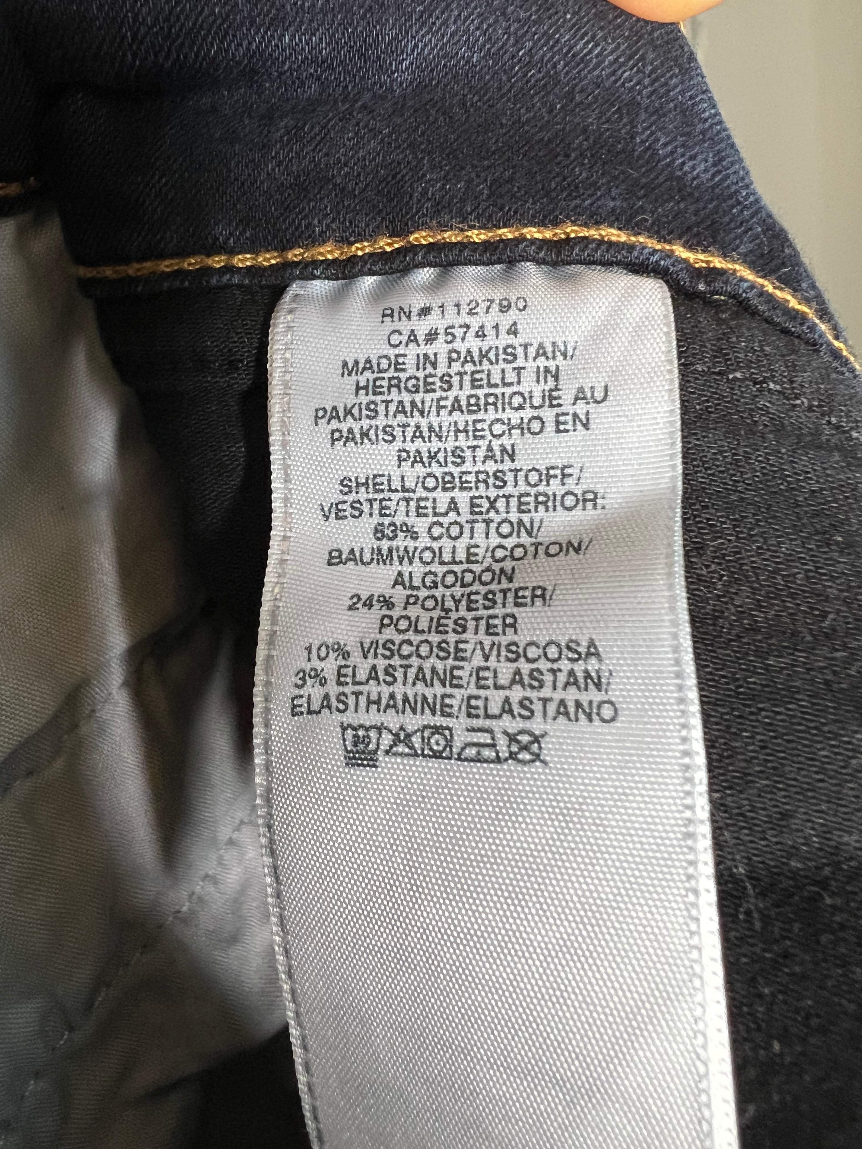 Close-up of a garment's interior label sewn onto dark blue denim fabric, showing text including 'MADE IN PAKISTAN' and care symbols on a light gray tag.