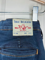Close-up of the back waistband of a pair of blue jeans hanging on a metal hanger with a plastic clip. The beige brand label reads 'True Religion' with a red Buddha graphic and 'World Tour' text.