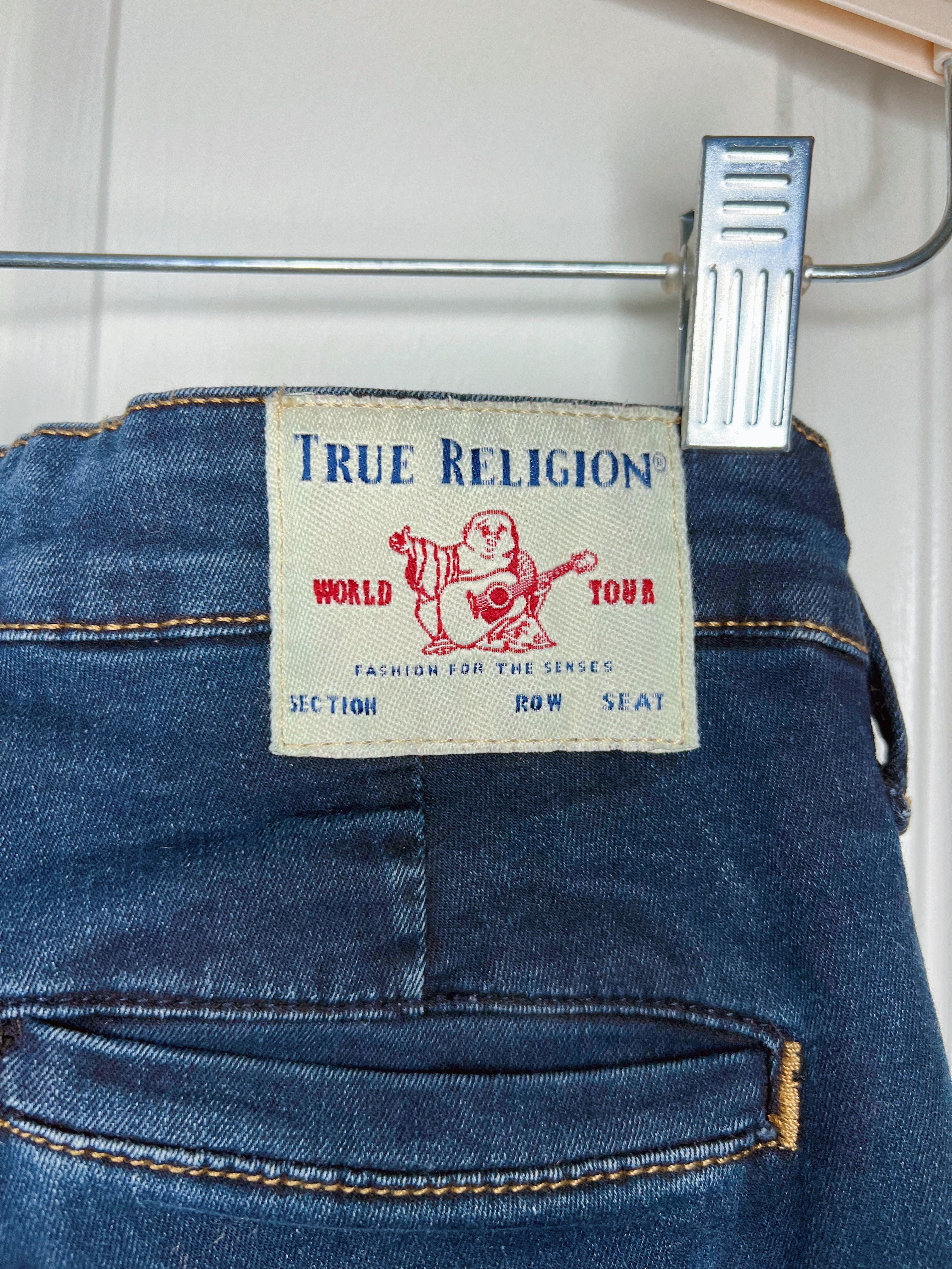 Close-up of the back waistband of a pair of blue jeans hanging on a metal hanger with a plastic clip. The beige brand label reads 'True Religion' with a red Buddha graphic and 'World Tour' text.