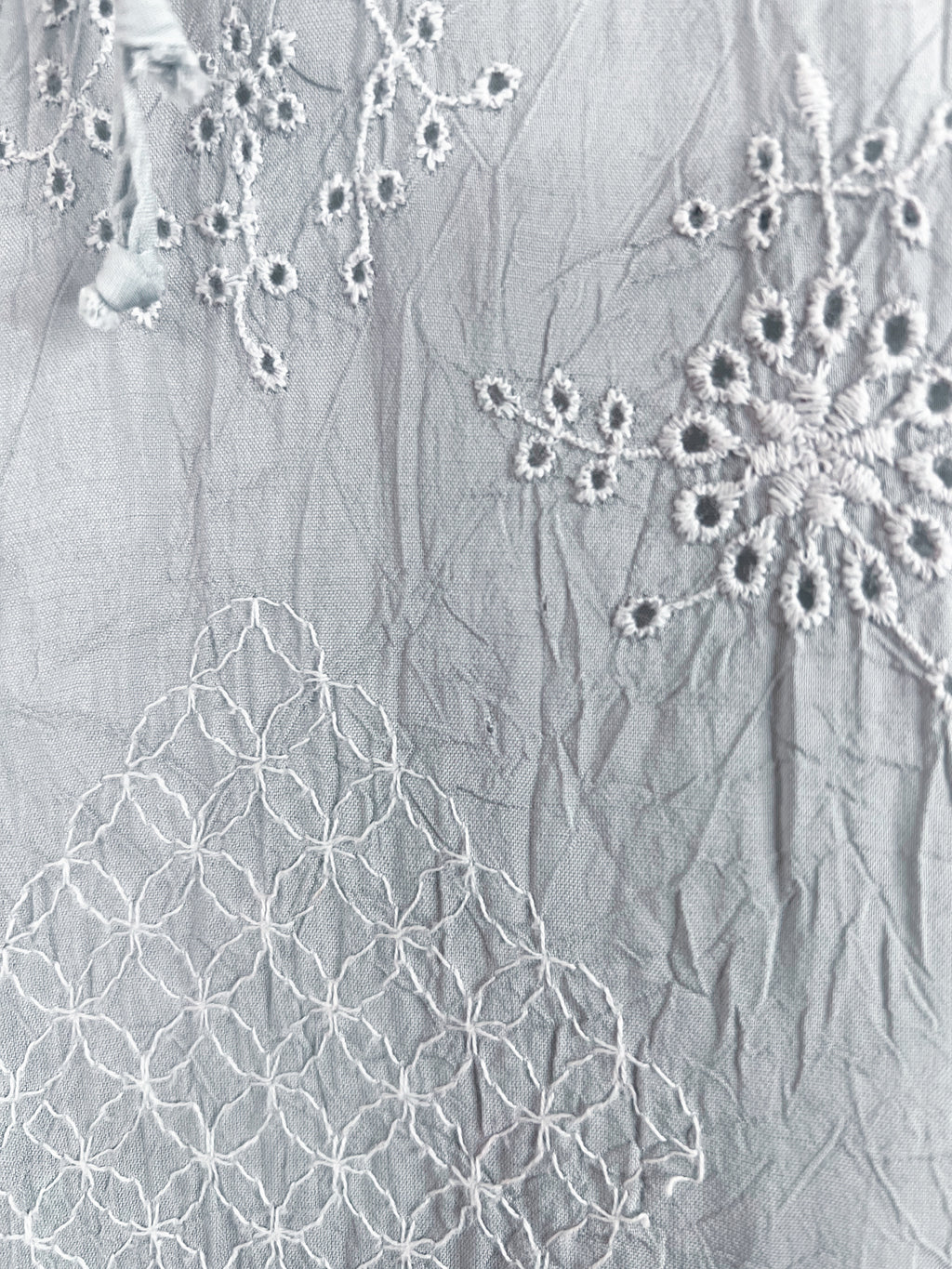A close-up of a light gray crinkled fabric with white embroidered floral and geometric patterns.