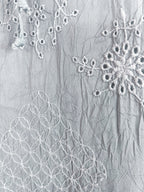 A close-up of a light gray crinkled fabric with white embroidered floral and geometric patterns.