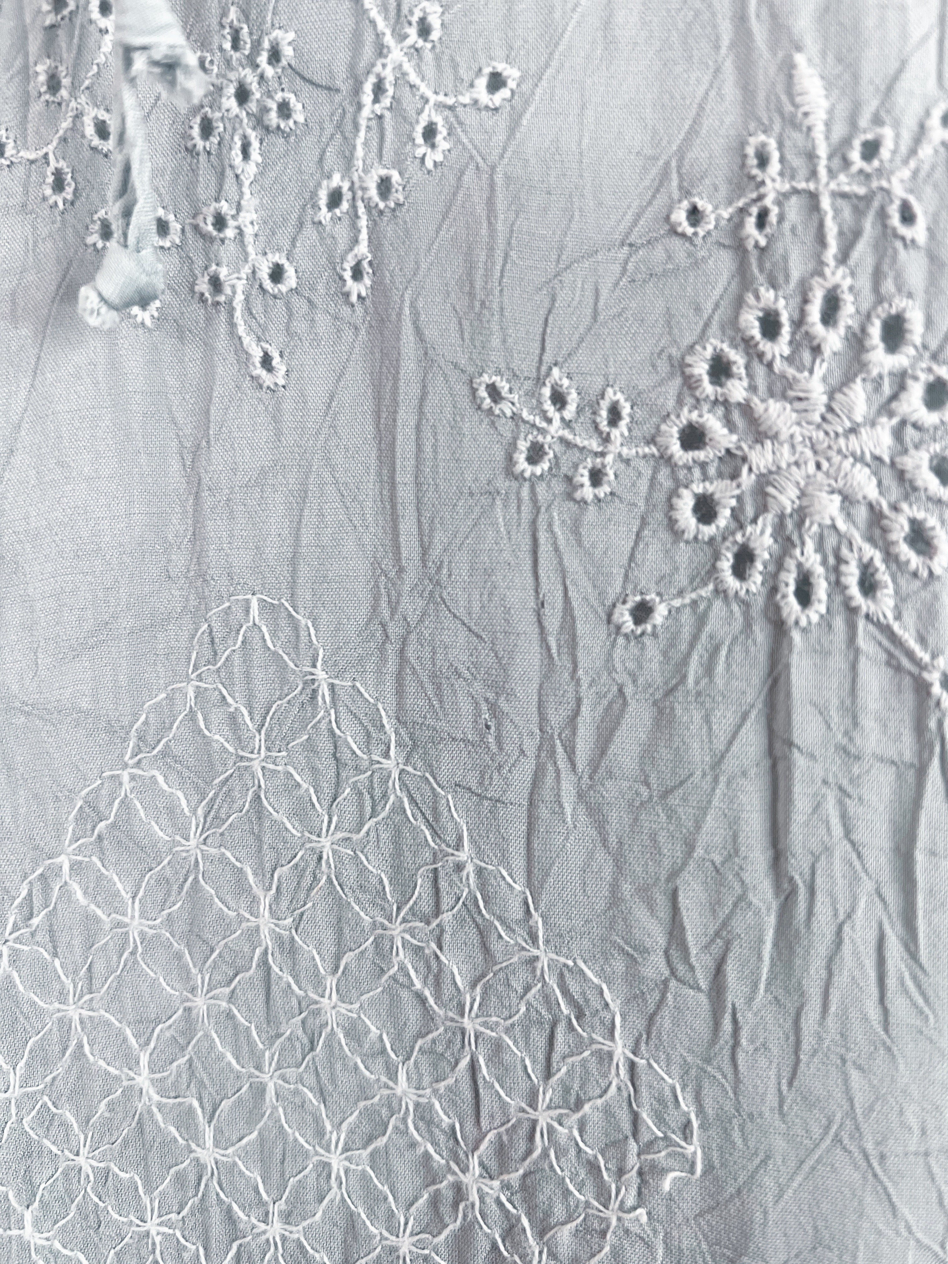 A close-up of a light gray crinkled fabric with white embroidered floral and geometric patterns.