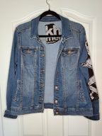 A blue denim jacket hanging on a black hanger against a white paneled door. The jacket features a black and white graphic print on the right sleeve and a black brand label on the inner collar.