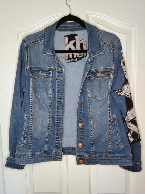 A blue denim jacket hanging on a black hanger against a white paneled door. The jacket features a black and white graphic print on the right sleeve and a black brand label on the inner collar.