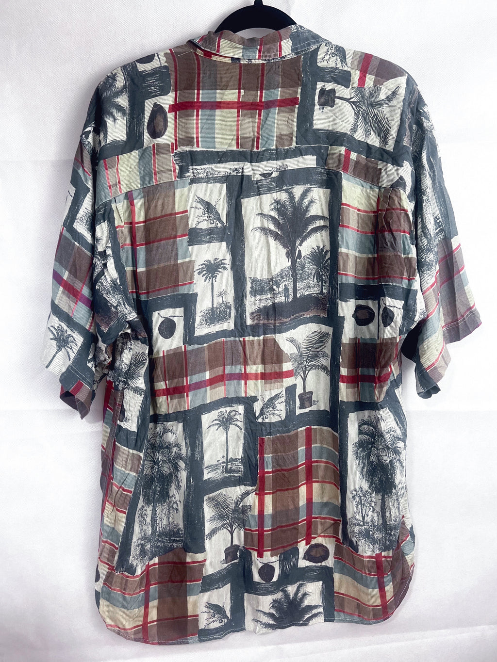 A short-sleeve button-up shirt with a patchwork print featuring plaid patterns, palm tree illustrations, and black-and-white tropical scenes on a white background.