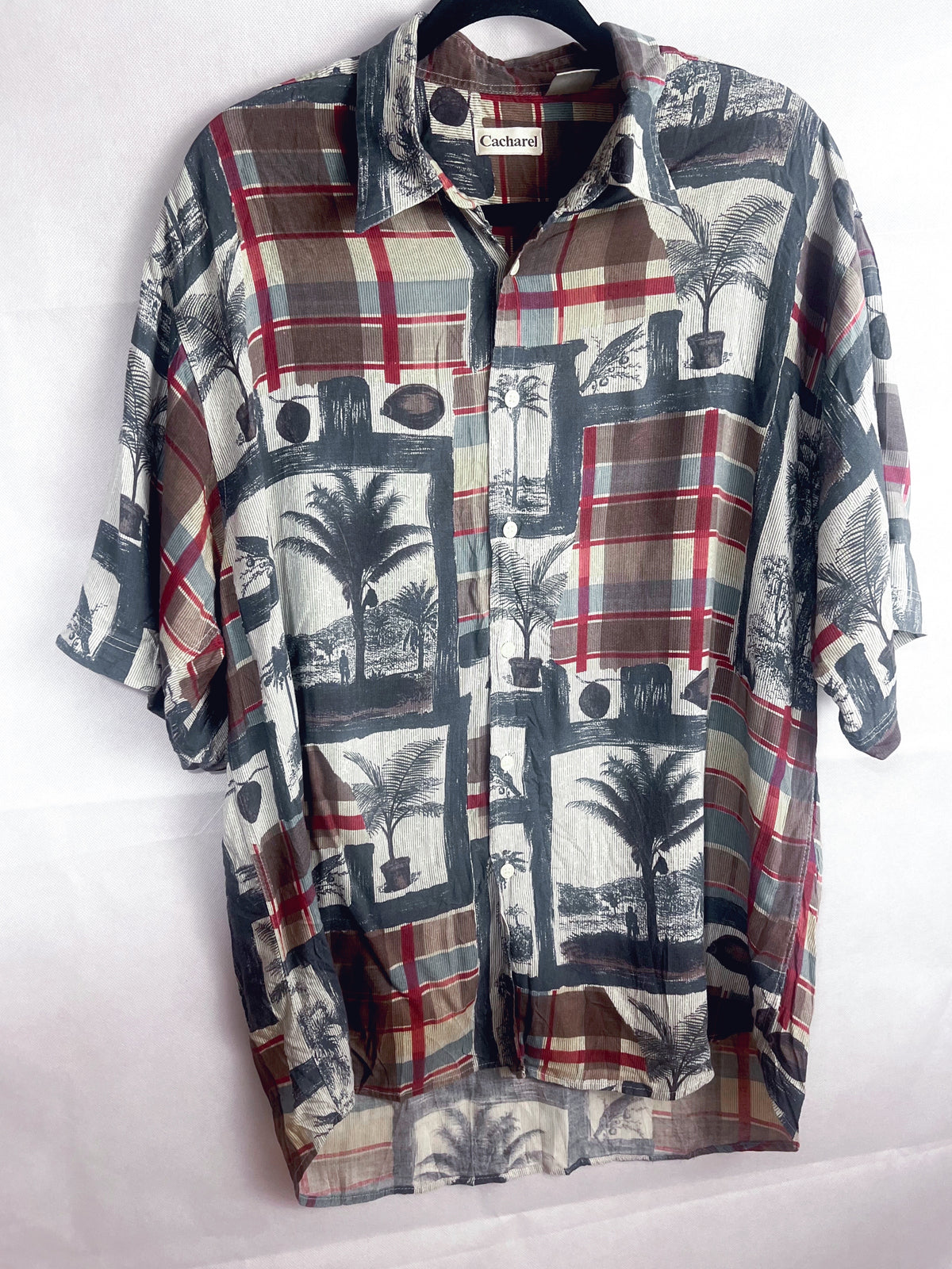 A short-sleeve button-up shirt with a mixed graphic print featuring plaid patterns, palm trees, and architectural elements in red, beige, gray, and black, hanging on a black hanger against a white background.