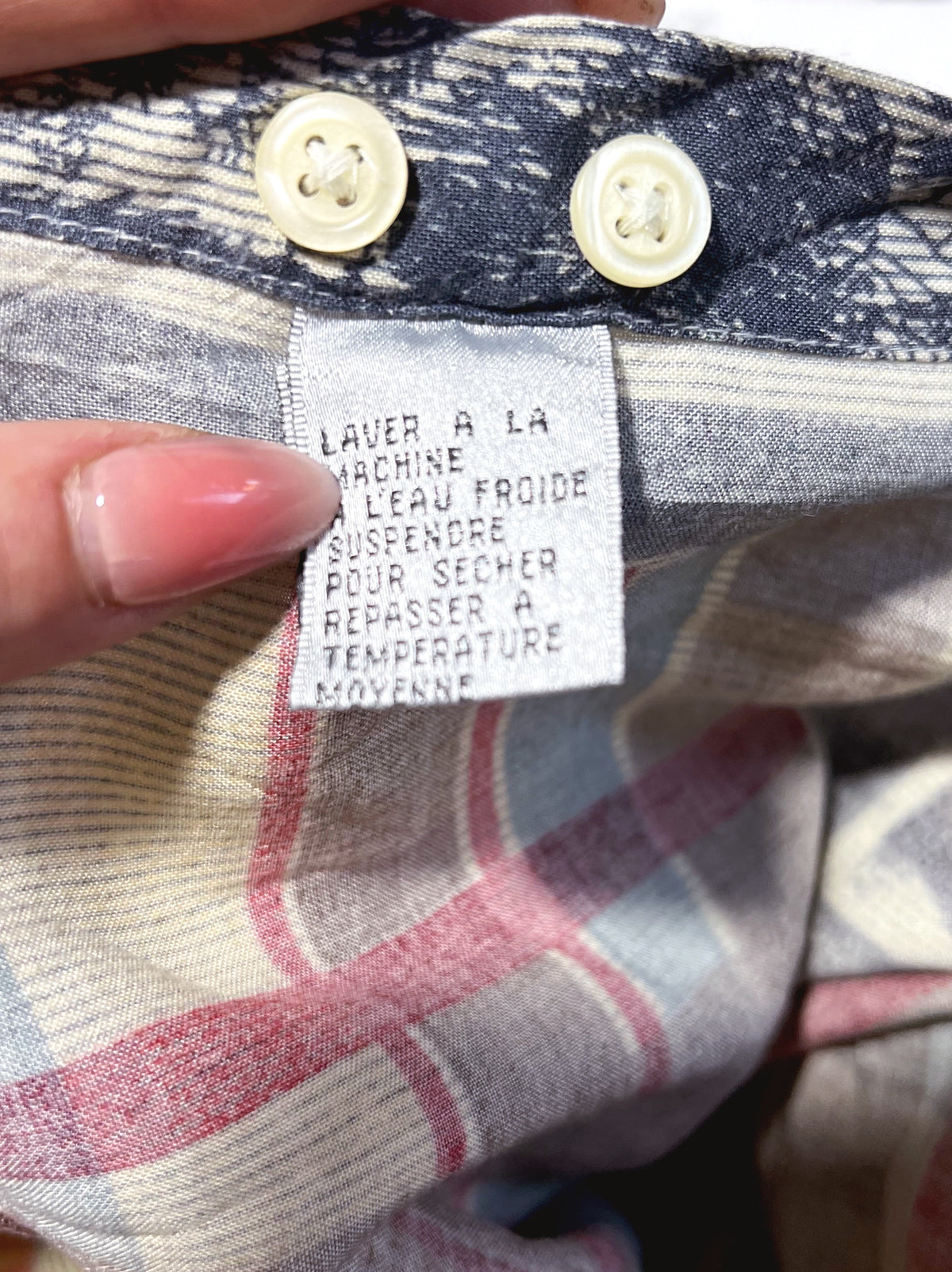 Close-up of a plaid shirt or garment fabric with a visible sewn-in care label and two cream buttons. A hand with pink nail polish is holding the fabric open.