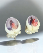 A pair of handmade shell-shaped earrings with white faux pearls at the base, featuring a translucent lavender and purple inlay with a pink and red floral design in the center, displayed on a reflective white surface.